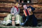 mężczyzna i kobieta w strojach regionalnych siedzą przed starym drewnianym domem, mężczyzna gra na akordeonie