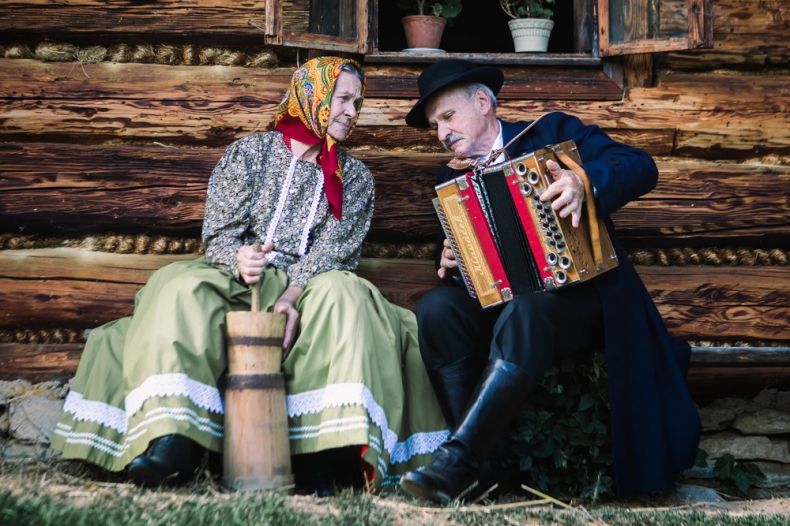 mężczyzna i kobieta w strojach regionalnych siedzą przed starym drewnianym domem, mężczyzna gra na akordeonie